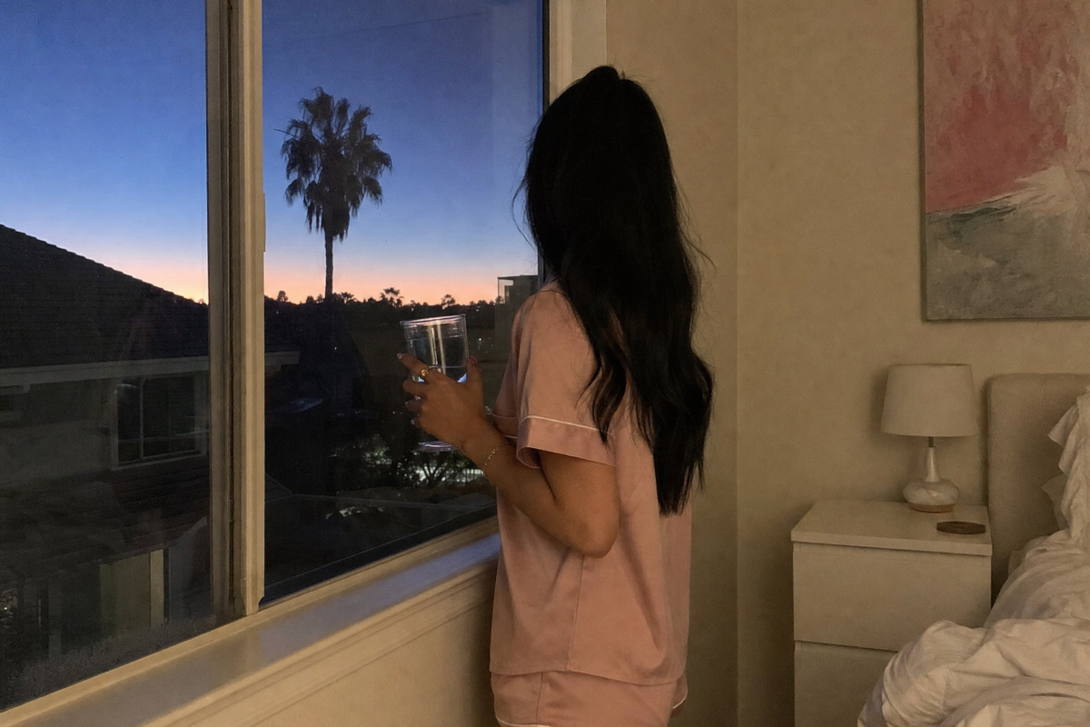 woman in a pink pajama set standing at a bedroom window at dusk holding a glass of water, looking out at a moonless sky