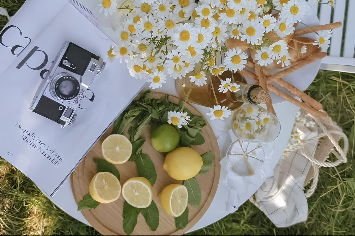Summer picnic flatlay with white daisies, halved lemons on a tray, a crochet bag, an open book, and a film camera - romanticizing everyday moments