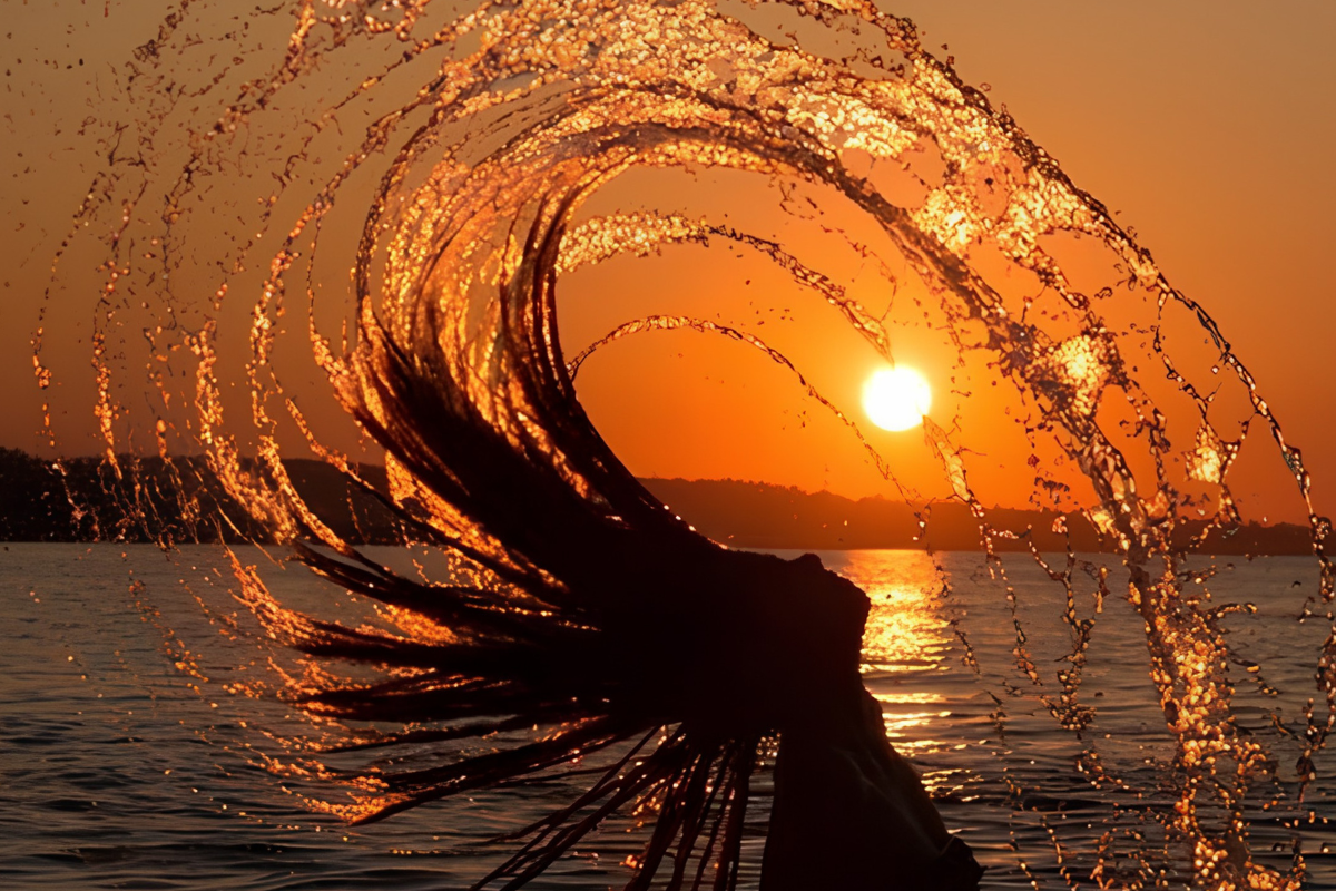 Woman flipping her hair in an arc of water against a golden sunset - celebrating the summer solstice and the start of Cancer season