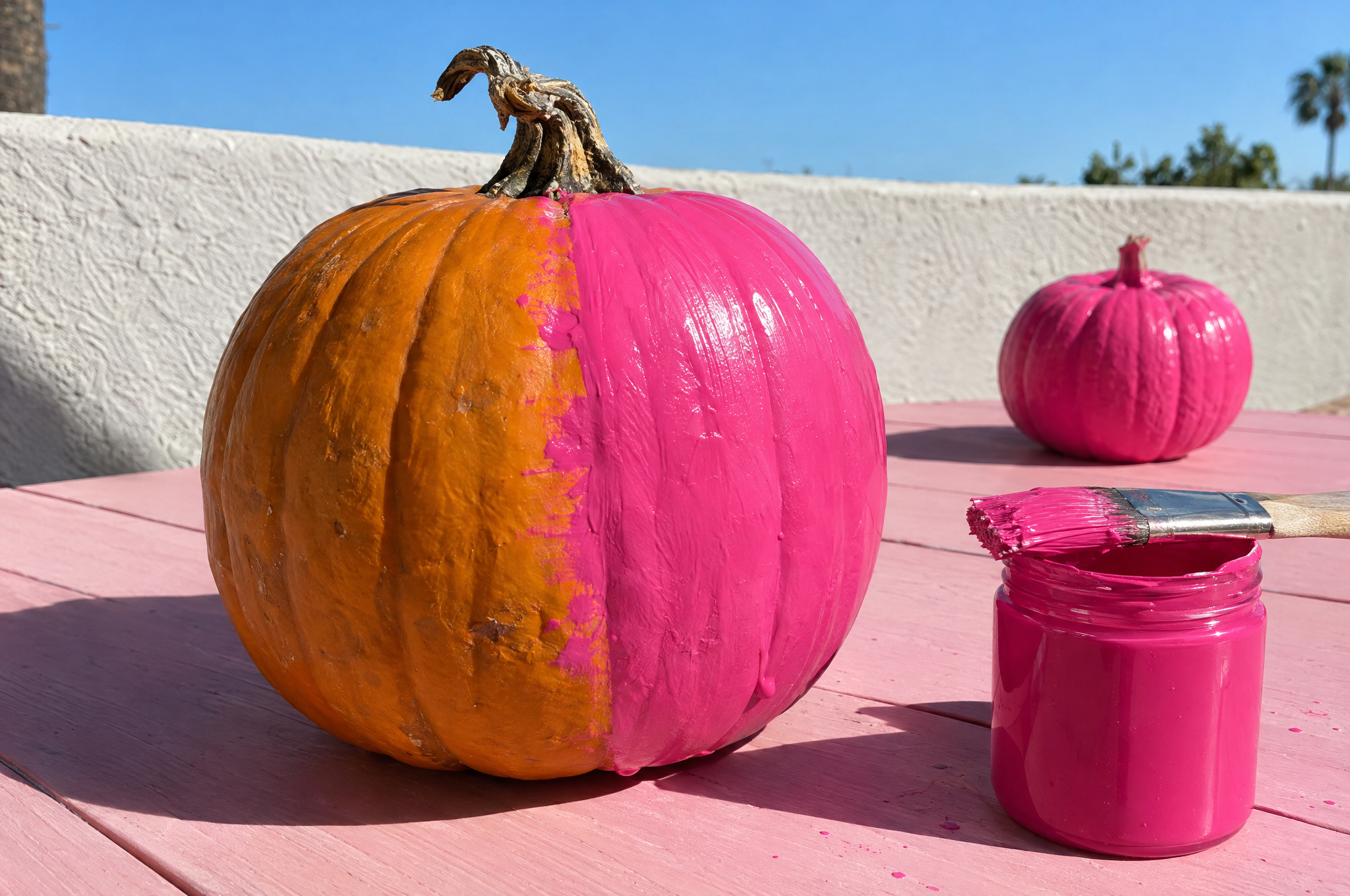 an orange pumpkin half painted hot pink beside a jar of pink paint and a brush, with a small pink pumpkin drying on a pink patio table under a blue october sky