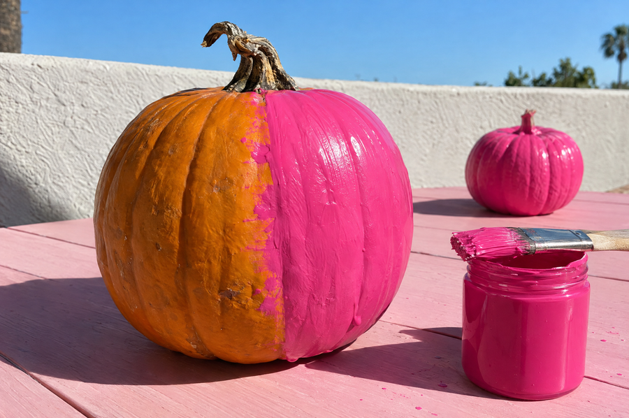 an orange pumpkin half painted hot pink beside a jar of pink paint and a brush, with a small pink pumpkin drying on a pink patio table under a blue october sky